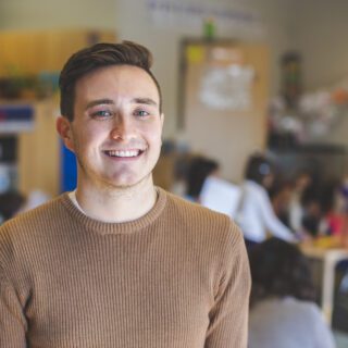 A young male teacher standing in a classroom with students in background behind him