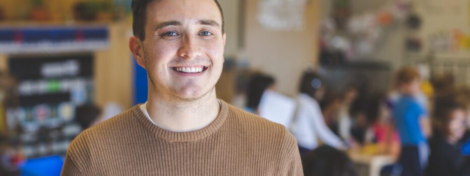 A young male teacher standing in a classroom with students in background behind him