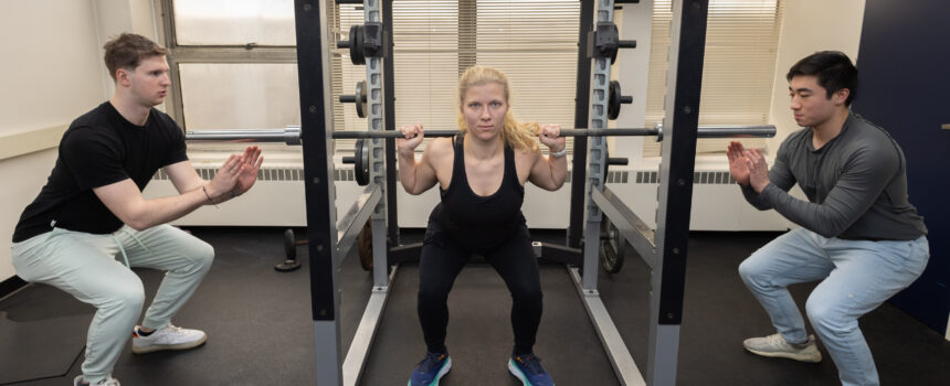 Student doing a squat with a weight bar as two students spot her on each side