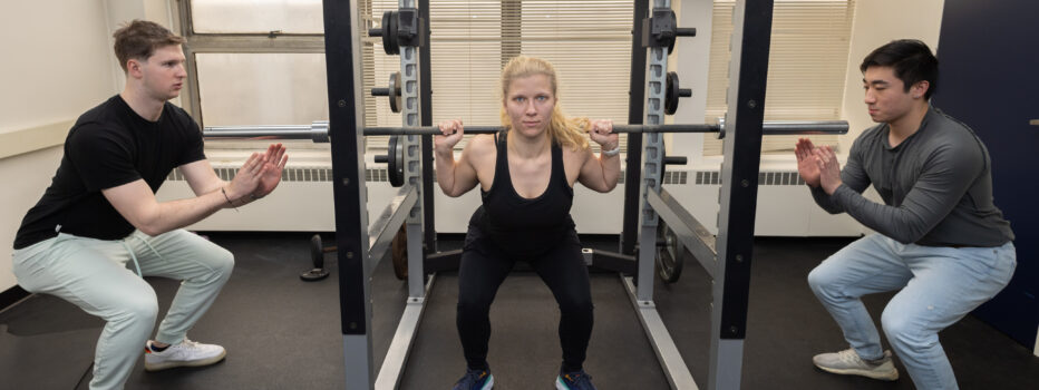 Student doing a squat with a weight bar as two students spot her on each side