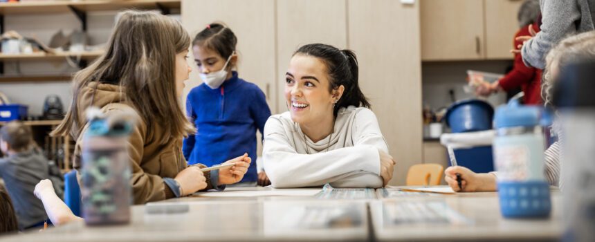 Student teacher crouches by the desk of a young student