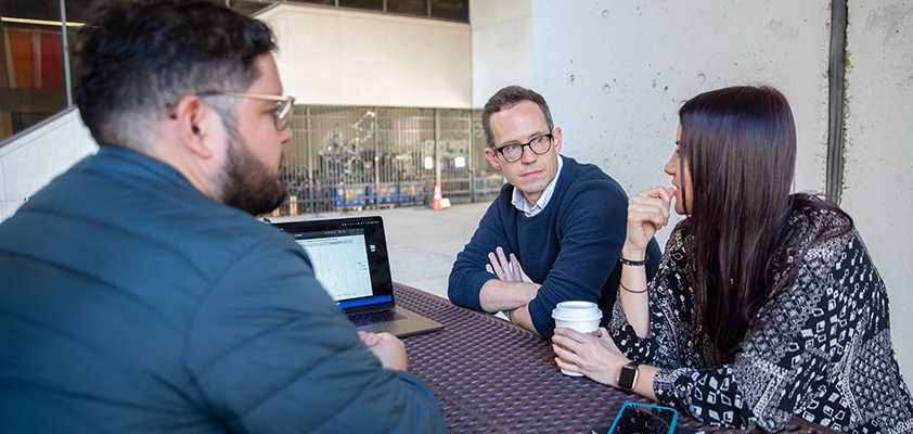 Three people with a laptop sit at a table outside Posvar Hall