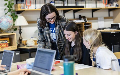 Student teacher looks over shoulders of two young students working at a desk