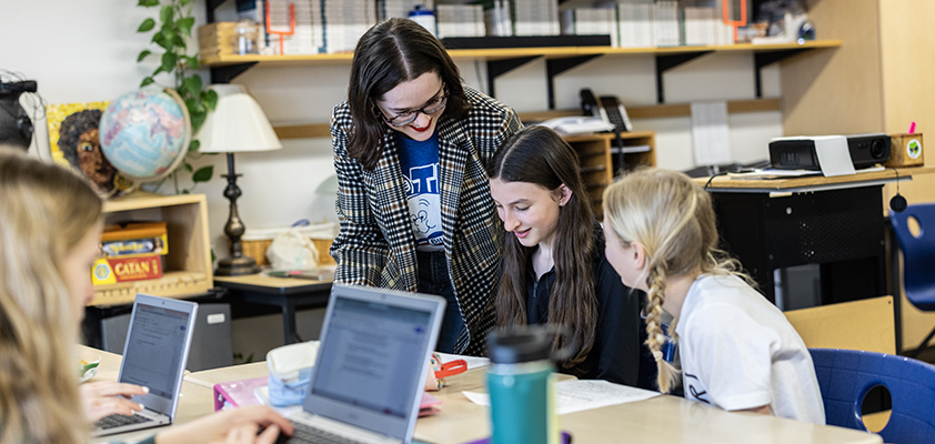 Student teacher looks over shoulders of two young students working at a desk