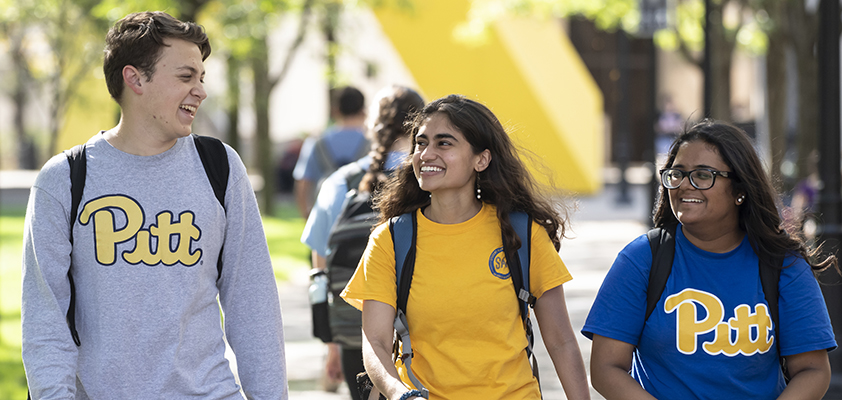 Three students in Pitt attire walk side-by-side on campus