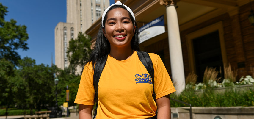 A female college student smiling and standing in front of the Cathedral of Learning on Pitt's campus