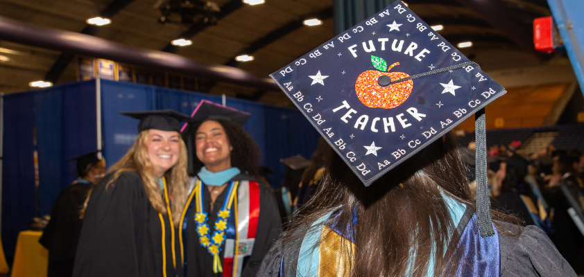 Graduation cap with "Future Teacher" design and two graduates in the background